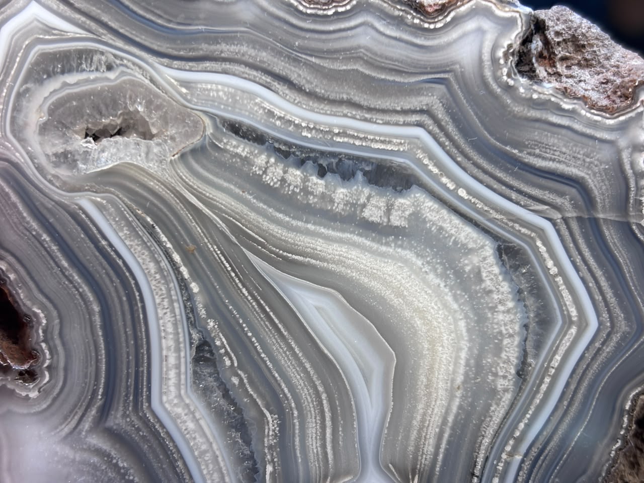 Close-up of a polished agate or marble-like stone slice with layered banding in white, gray, beige, and soft blue-gray tones, with darker brown edges.